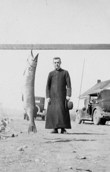 A reverend poses next to a large pike that hangs from a beam.