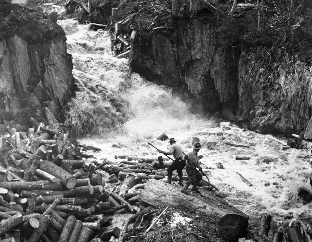 Two drivers carefully balance themselves while steering logs at the bottom of a waterfall.