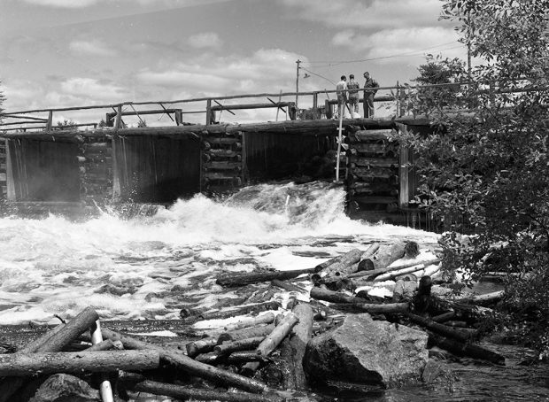 Three men are standing on a dam made of logs. A gate in the dam is open, allowing a torrent of water and logs to pass through.
