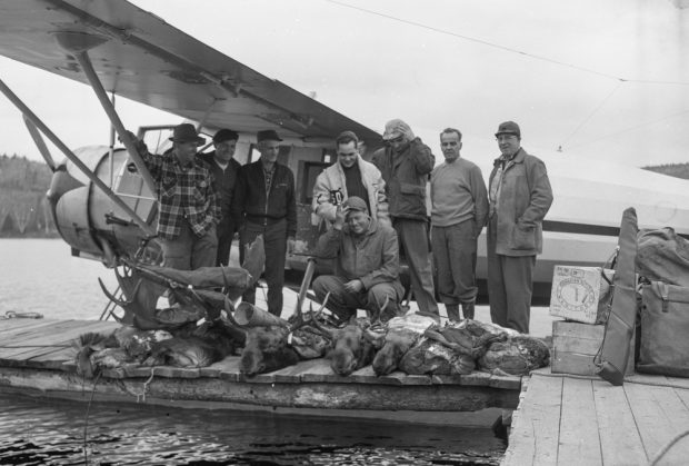 Eight men pose on a dock in front of a seaplane with four moose carcasses.