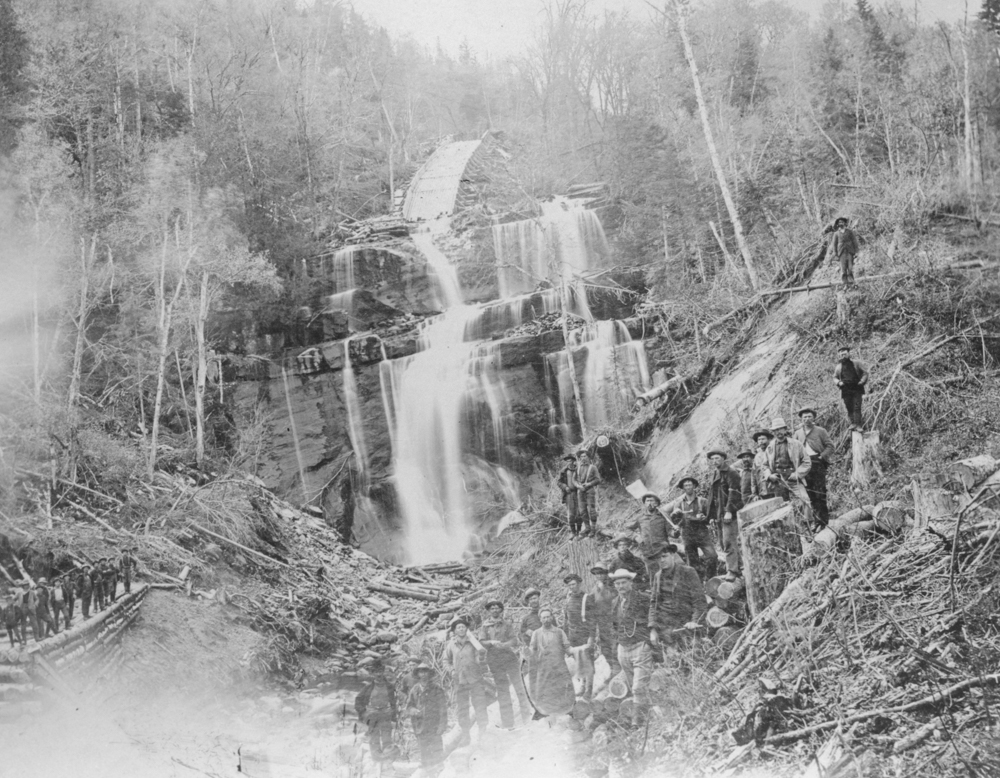 Plusieurs travailleurs posent au bas d'une chute d'eau où ils ont aménagé une rampe pour les billots de bois.