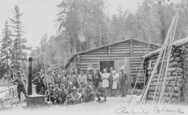 A group of workers pose in front of small log cabins in the middle of the forest.