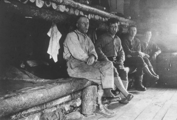 Men are sitting on beds made of fir trees and rough-cut logs.