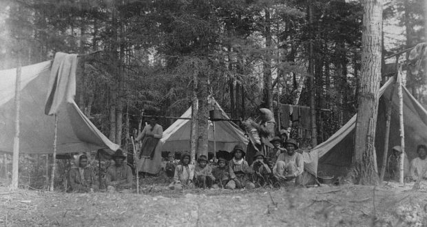 Indigenous people of all ages pose in the midst of rudimentary tents surrounded by tall coniferous trees.