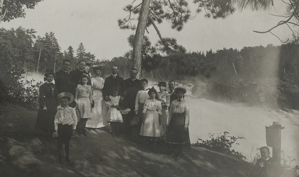 15 people, including women, children and clergy, pose at the top of the falls.