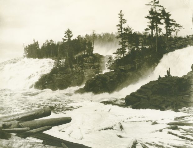 The water falls violently down the Shawinigan Falls, divided into three sections, through rocks and piles of logs.