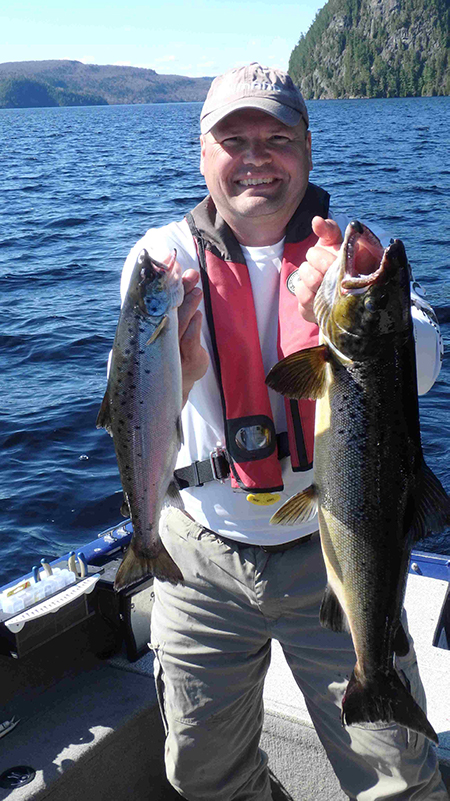 Mario Genois, tout sourire dans une chaloupe au milieu de la rivière, tient deux poissons par les branchies.