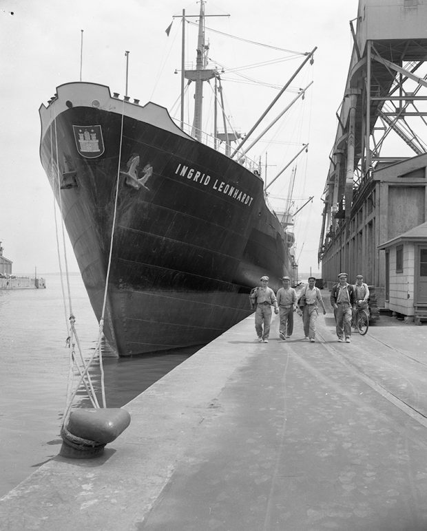 Men walking on a quay in front of the huge bow of a docked ship.