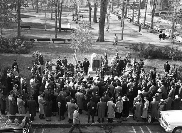 A crowd is gathered near a park. People are gathered in front of a large rock on which a bronze plaque is placed. A man addresses the crowd in front of microphones.