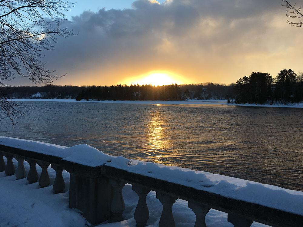 The sunset is reflected on the river water. A snow-covered railing is in the foreground.