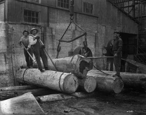 In front of the factory, three men are standing on large trunks that are tied together with chains.