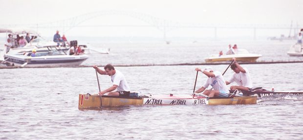 Two men are rowing boldly in a canoe. Pleasure crafts are watching them in the background. Another canoe follows closely behind.