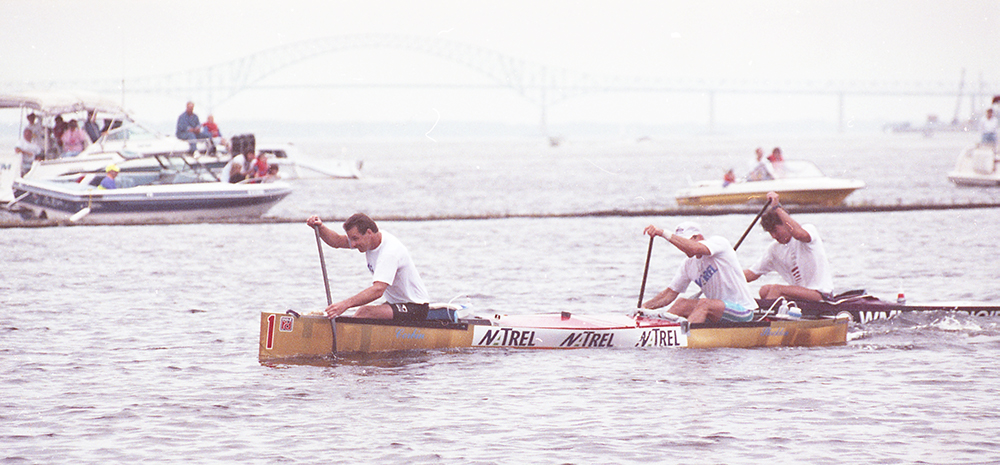 Deux hommes rament hardiment à bord d’un canot. Des bateaux de plaisance les regardent en arrière plan. Un autre canot les talonne de près.
