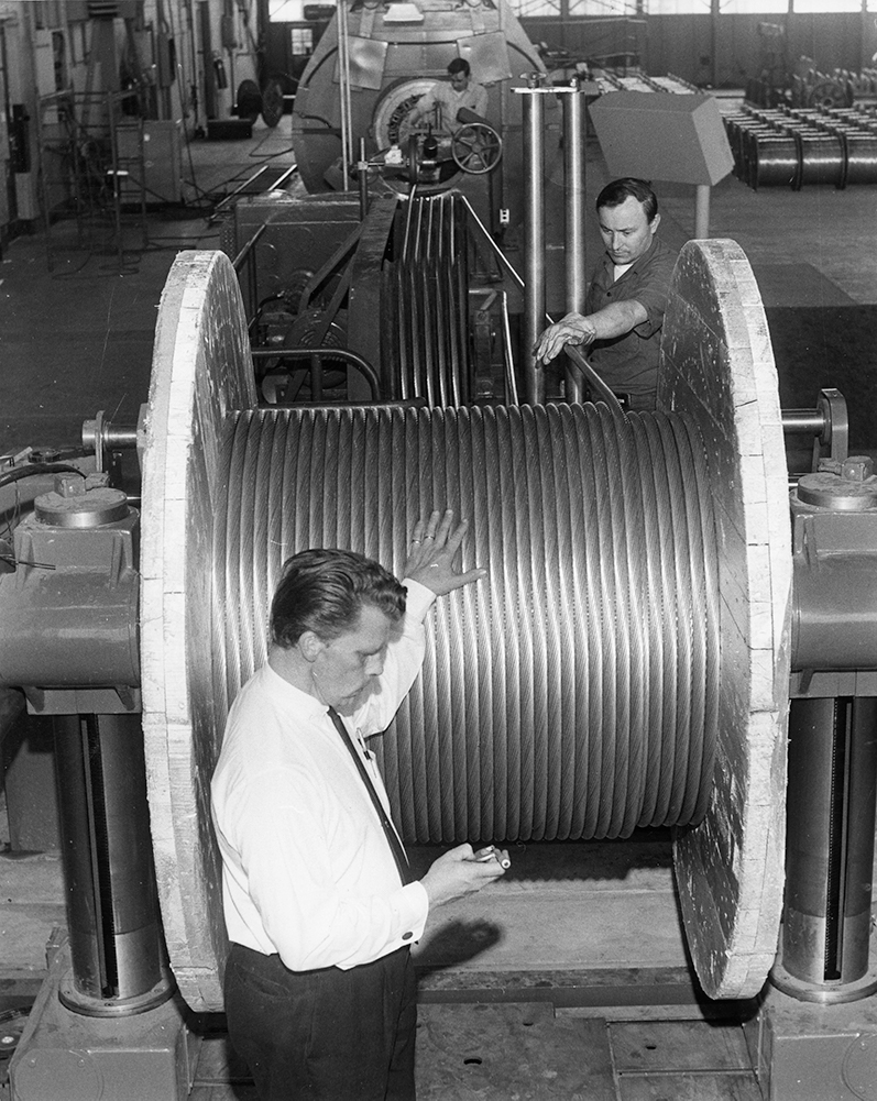 Employees monitor the winding of a large aluminum cable onto a large wooden spool.