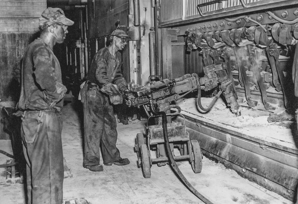 Two employees, wearing wool clothing, caps and small glasses, are handling machinery near an aluminum pot.