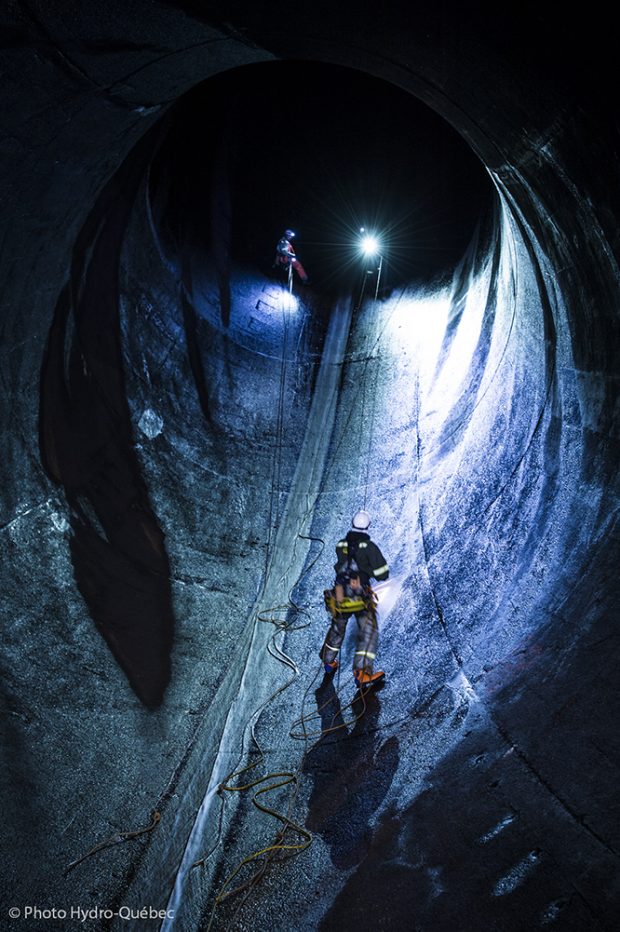 A worker with climbing equipment stands in the light of a searchlight inside a huge concrete tunnel, which functions as a penstock.