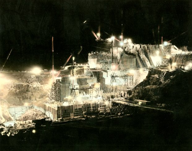 Site of a dam under construction. The huge cranes and scaffolding are lit by searchlights.