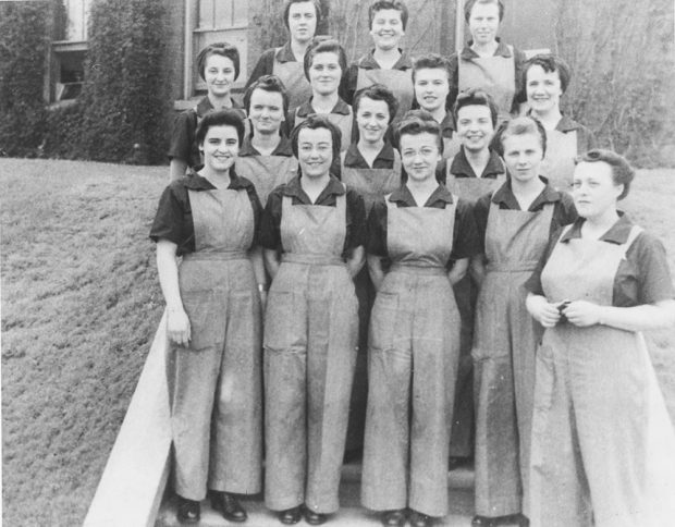 A group of women in work clothes pose in front of a brick building.