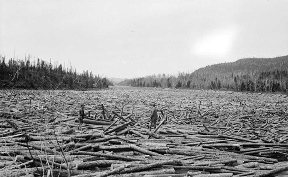 Two men stand on logs that cover the entire river