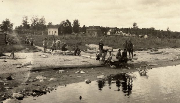 A group of people are standing on a beach. Behind them are modest wooden houses across the land.