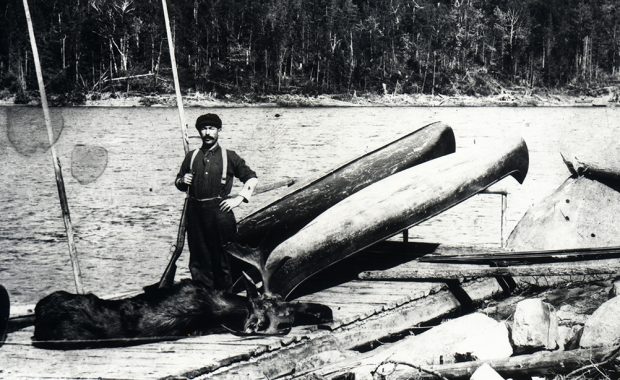 A man poses with a moose carcass on a dock in front of a lake.