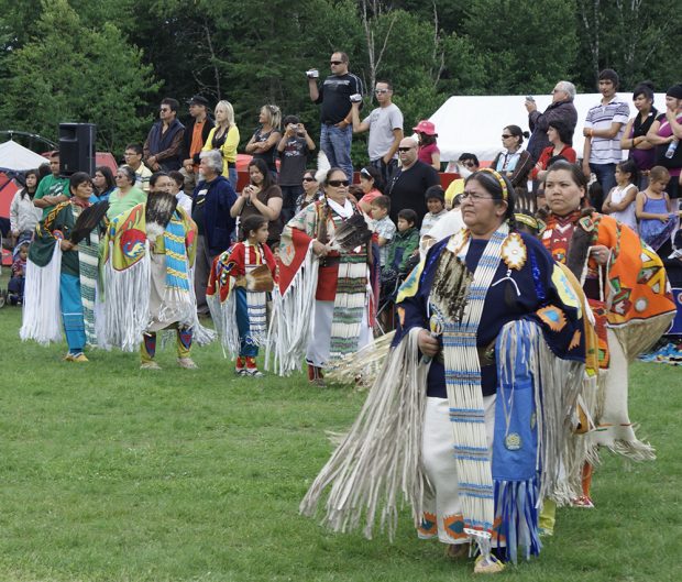 Indigenous people in traditional dress perform a traditional dance in front of packed bleachers.