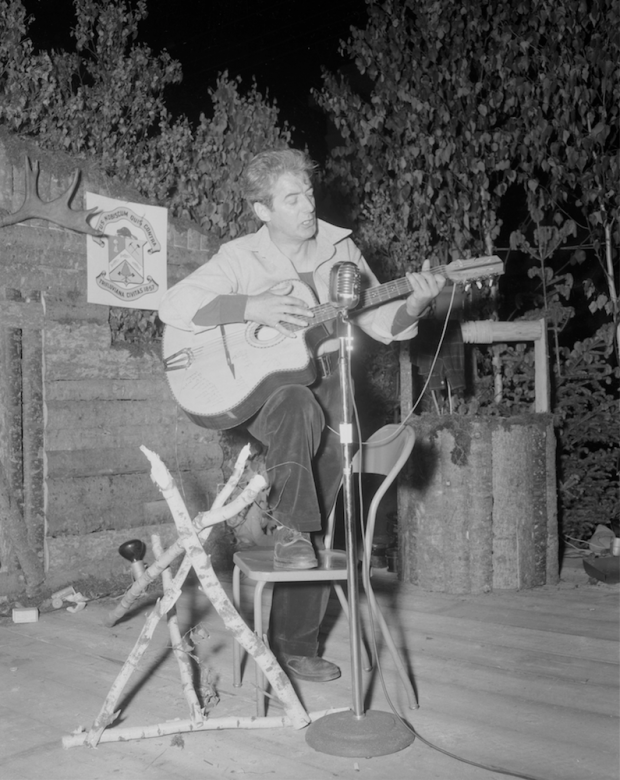Félix Leclerc sings and plays guitar on a stage decorated with logs