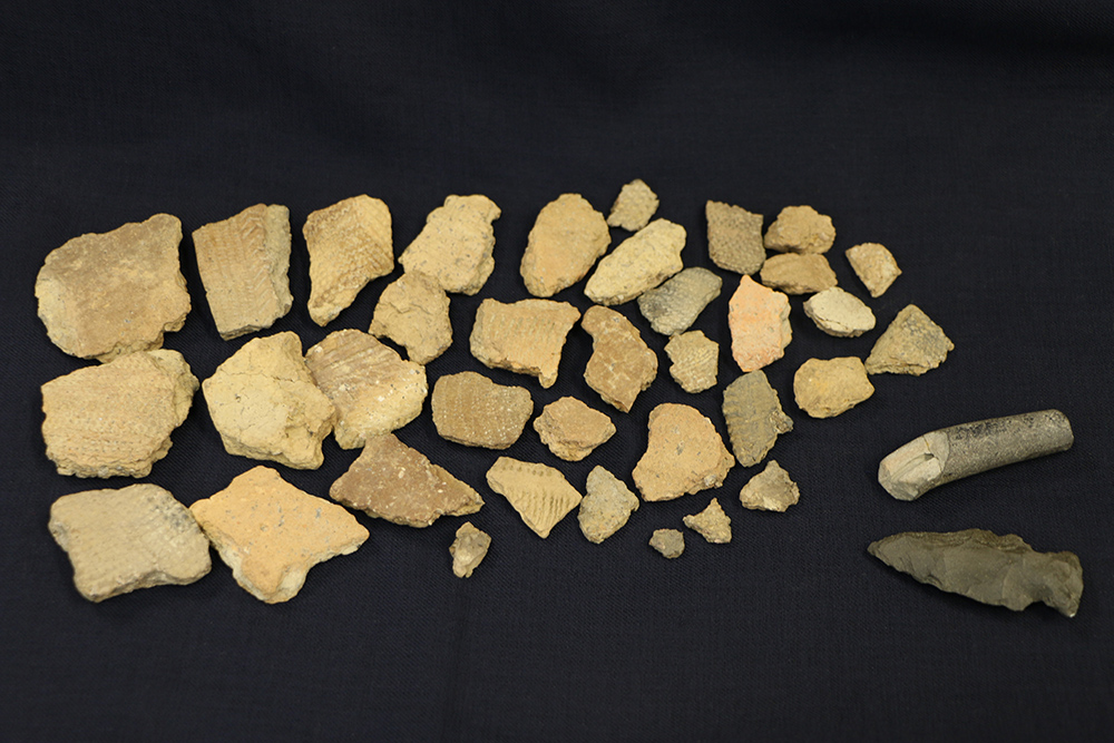 Artefacts amérindiens disposés sur un fond noir. Au bas à droite, une pointe de projectile en forme de flèche et un fragment de pipe en céramique, de couleur grise. À gauche, une vingtaine de tessons de poterie, beige et gris ornés de motifs en chevrons.