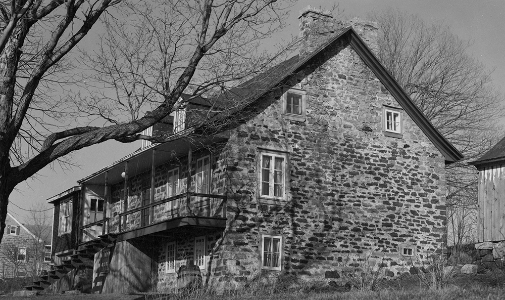 Black and white photograph of an 18th-century stone and wood house, side view. The house has solid stone chimneys and a large wooden porch.