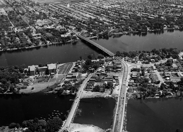 Black and white aerial view of a bridge spanning a wide river. There are neighbourhoods on both shores.