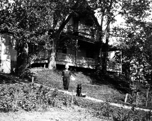 Black and white photograph of a woman standing with her dog on a road in front of a large house in summer.