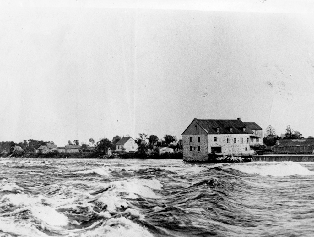 Black and white photograph of a mill in the distance. A river flows in the foreground. The waves are rough.