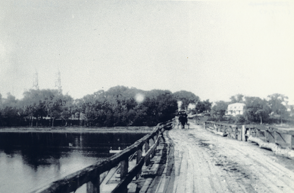 Black and white photograph of the Bellefeuille Bridge. The wooden bridge, with railings on the sides, is not straight. A sled can be seen at the far end.