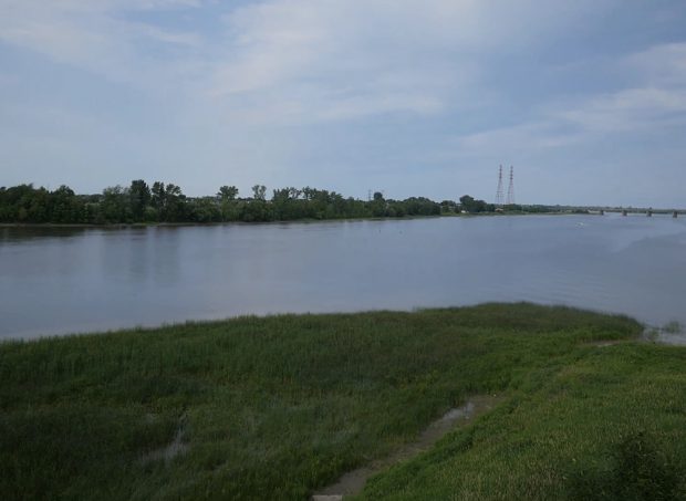 The video begins above the shores of the eastern portion of Île Jésus in summer. The trees and grass are dark green. The camera rises gradually above the trees. The Mille Îles River appears. The camera turns to focus on the area where the two rivers meet. In the distance, you can see the Charles De Gaulle Bridge in Repentigny and the St. Lawrence River. A boat is moving in the water and cars are travelling on the bridge.