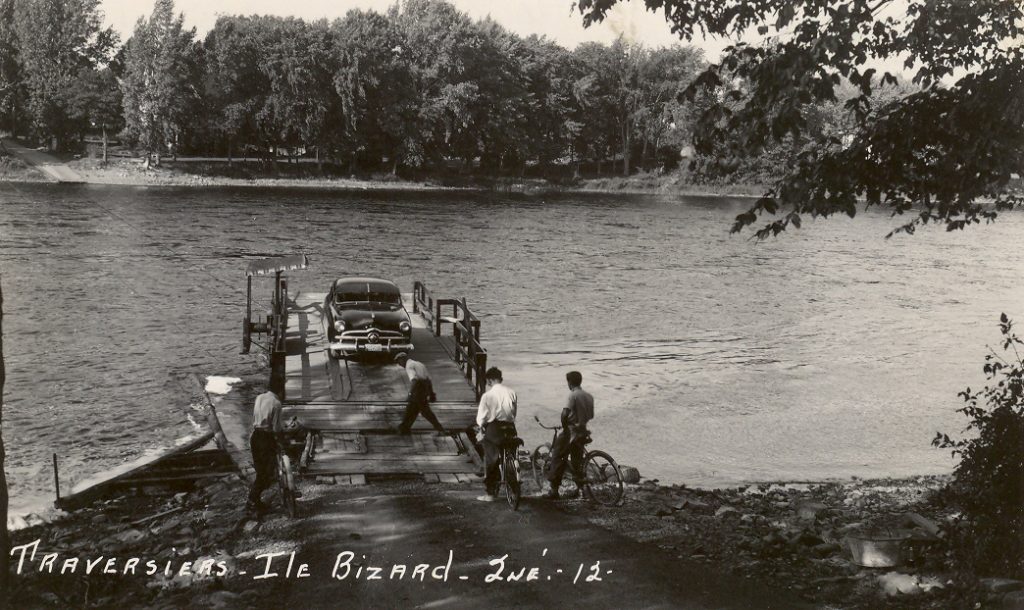 Photographie en noir et blanc. Une voiture est installée sur la plateforme de la traverse. Des hommes à vélo attendent leur tour. La rive de l’île Bizard est en arrière-plan.