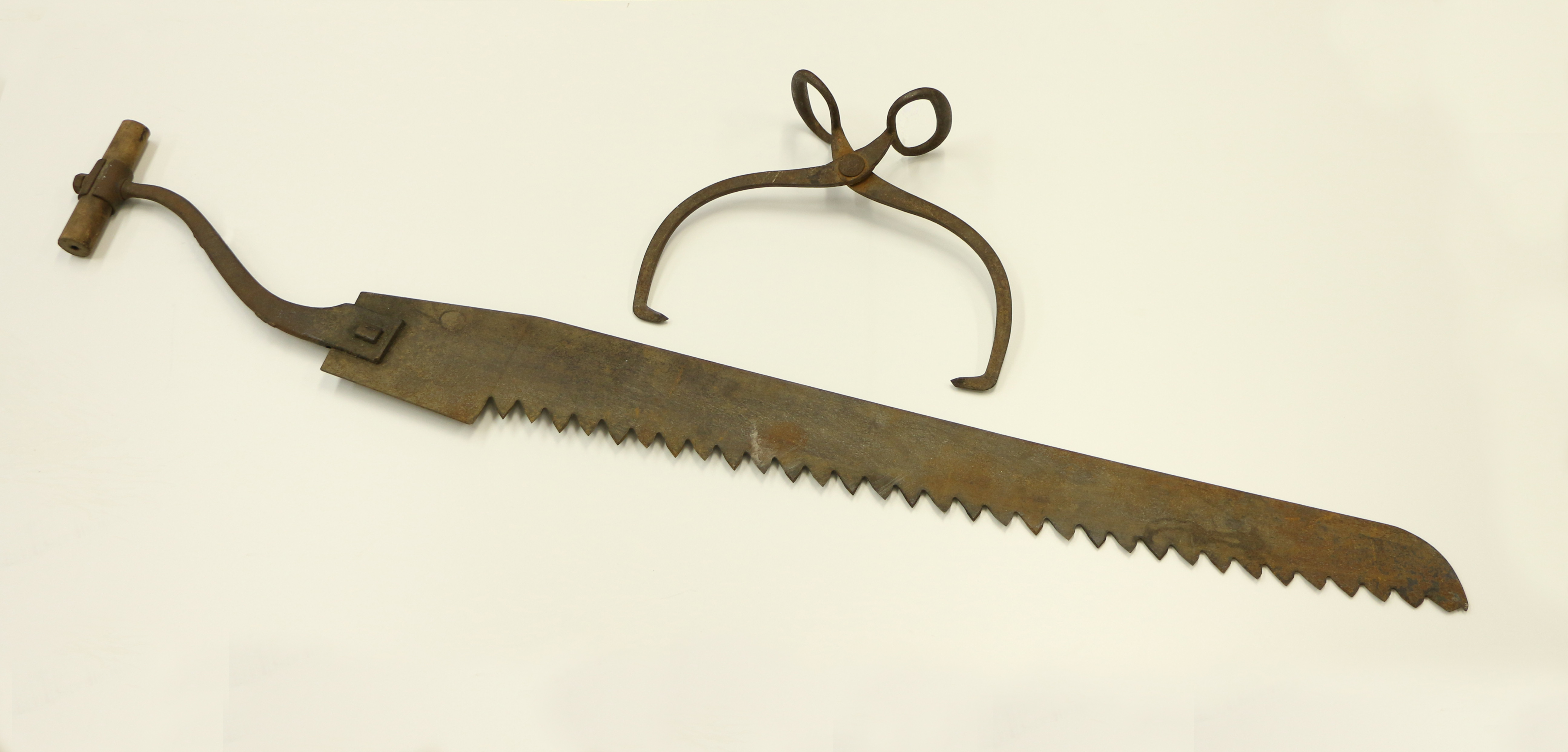 Photograph of ice-cutting tools. On a white background, brown metal ice tongs used to carry blocks of ice, and a rusted iron saw with a wooden handle.