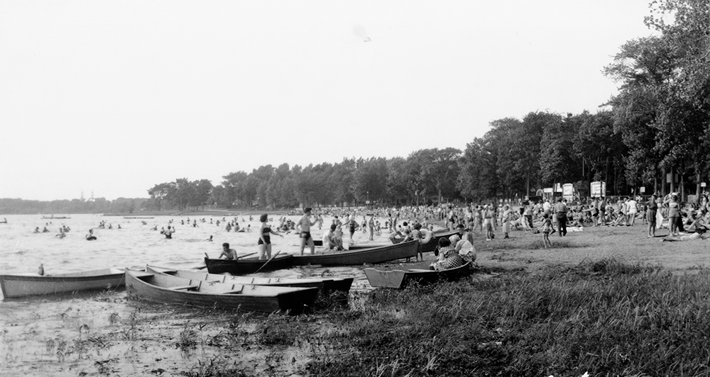 Black and white photograph of a beach with boats and lots of bathers. The beach is surrounded by many trees.