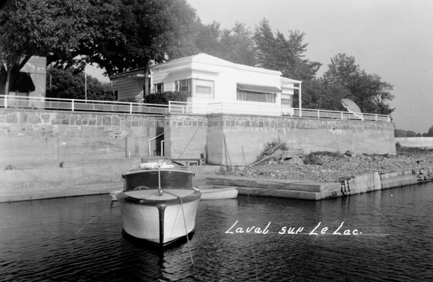 Black and white photograph of a white house with a small pleasure boat tied up at a grey stone dock.