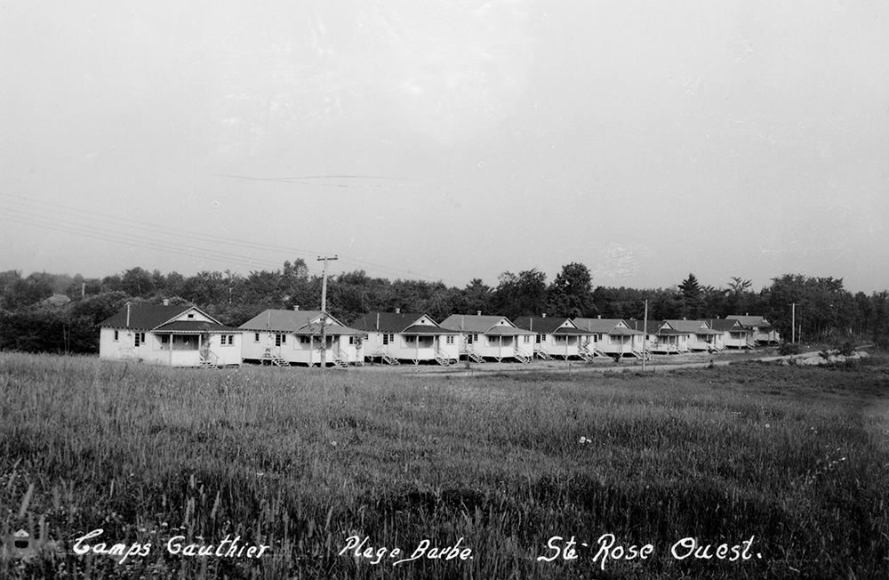 Photographie en noir et blanc d’un regroupement de 10 petits chalets identiques en bois blanc, construit un à la suite de l’autre.