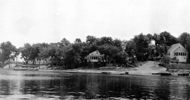 Black and white photograph of three wooden cottages surrounded by trees on a river.