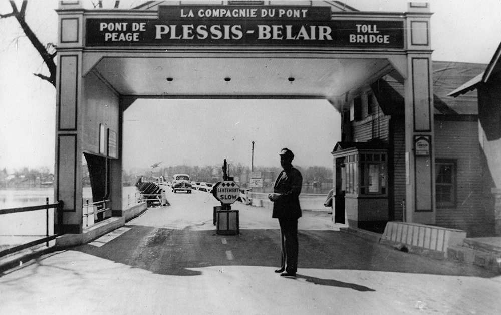 Black and white photograph of the entrance to the Plessis-Bélair Bridge. The name of the bridge and the words “pont à péage / toll bridge” are written on an arch. A security guard stands in front of the toll booth.