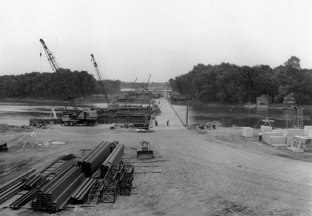 Photographie en noir et blanc de la construction de l’autoroute 15. Deux travailleurs, une grue et des matériaux de construction sont visibles, ainsi que des grosses poutres de métal et des morceaux de bois.