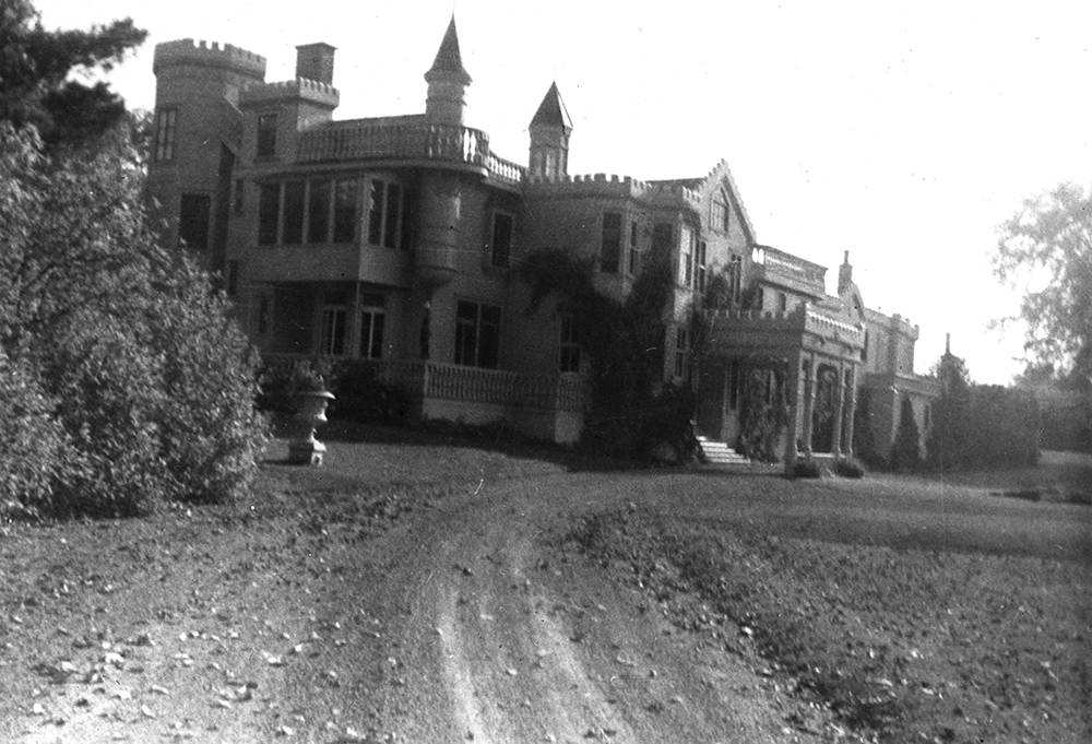 Photographie en noir et blanc d’un manoir de grande taille et d’un chemin de terre qui mène à la demeure. Le manoir a une toiture irrégulière avec tourelles et pignons.