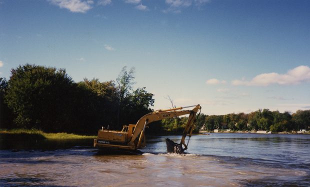 Colour photograph of a yellow mechanical shovel doing clean-up work on the Des Prairies River on a sunny summer day.