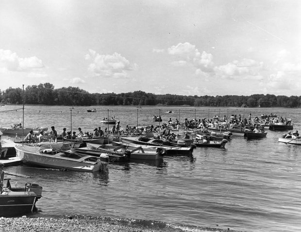 Black and white photograph of small motorboats moored to a dock. Many people are sitting in them, ready for a boat parade.