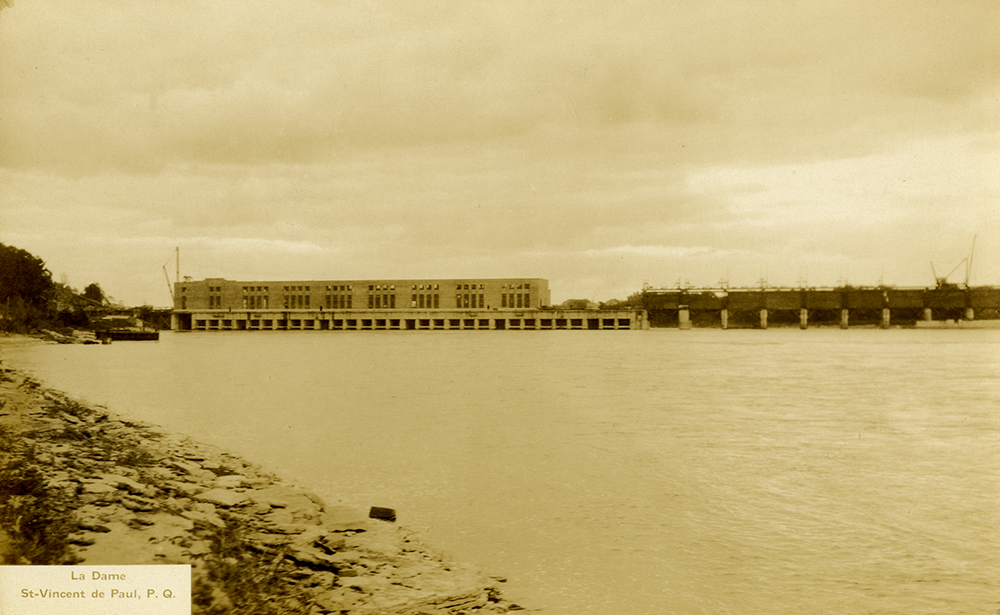 Yellowed photograph of the construction of a dam, taken from shore. The large dam across the Des Prairies River can be seen in the distance.