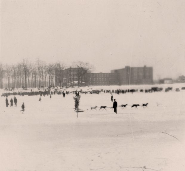 Black and white photograph of a dogsled race on the Des Prairies River in winter. Hundreds of people are standing on the river’s snowy shore and six dogs harnessed together are pulling a sled.