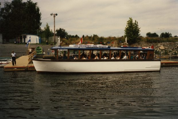 Colour photograph of a white boat moored and filled with passengers ready for a cruise on the Des Prairies River.