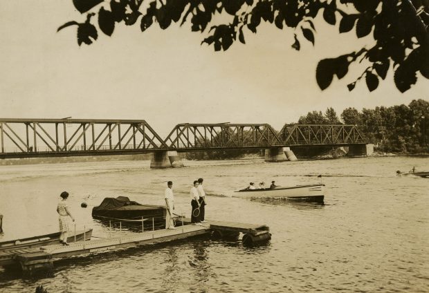 Sepia photograph of people standing on a dock looking at motorboats on the river. In the background, a train bridge spans the river.