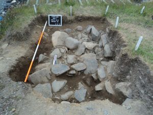 Grid parameters and scale lying next to pile of stone rubble in a rectangular excavation.